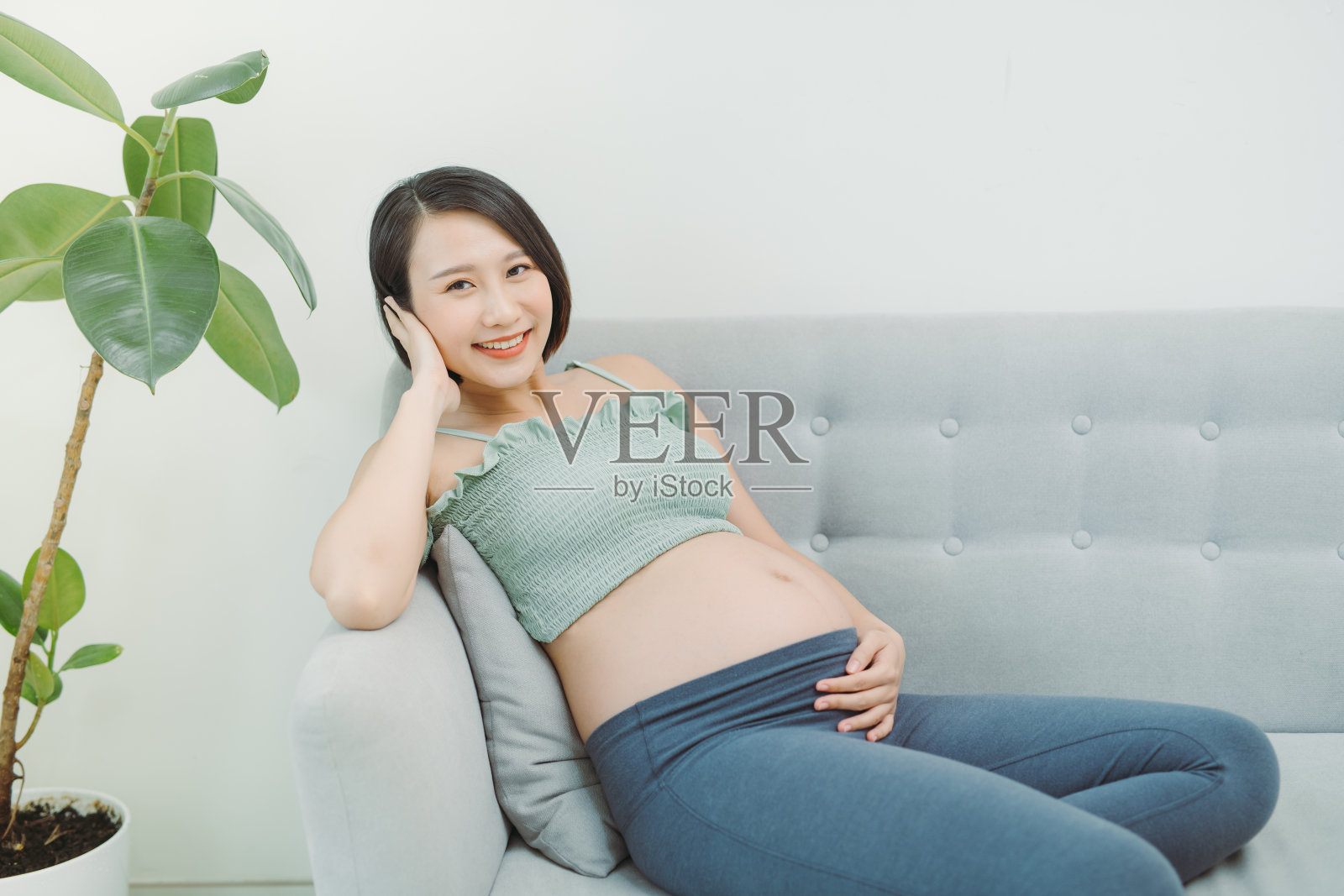 怀孕快乐微笑的女人坐在沙发上，爱抚她的肚子。照片摄影图片