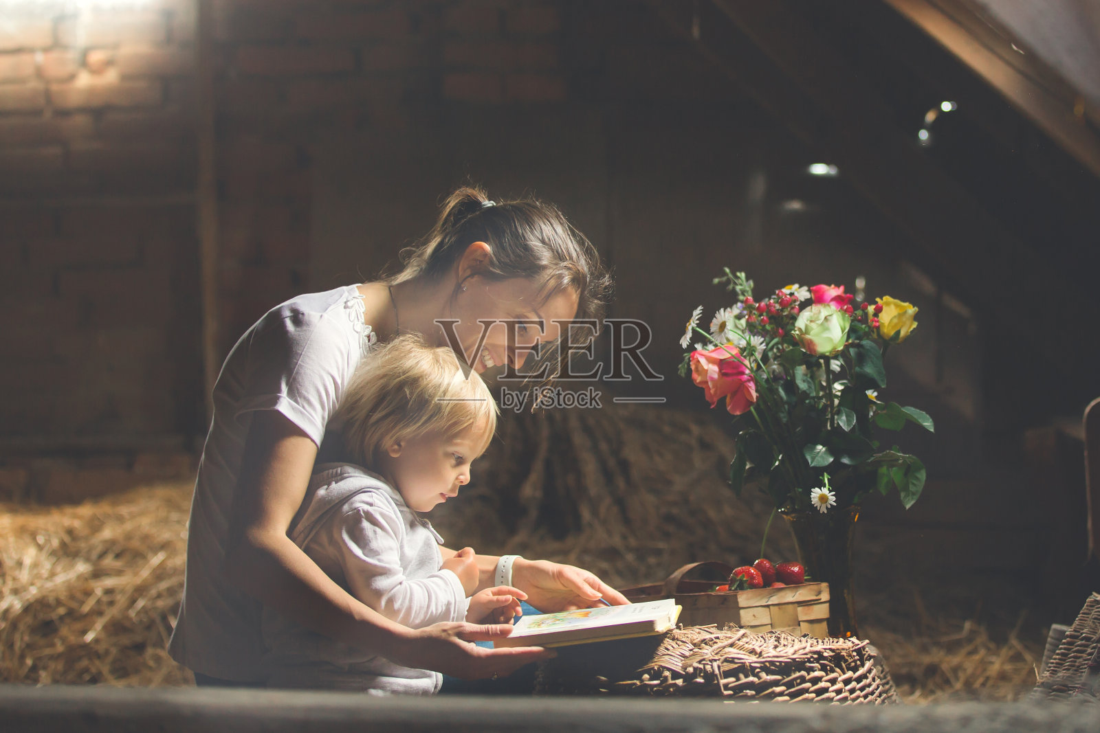 漂亮的妈妈和蹒跚学步的金发男孩，在阁楼上看书，不错的氛围，鲜花，草莓照片摄影图片
