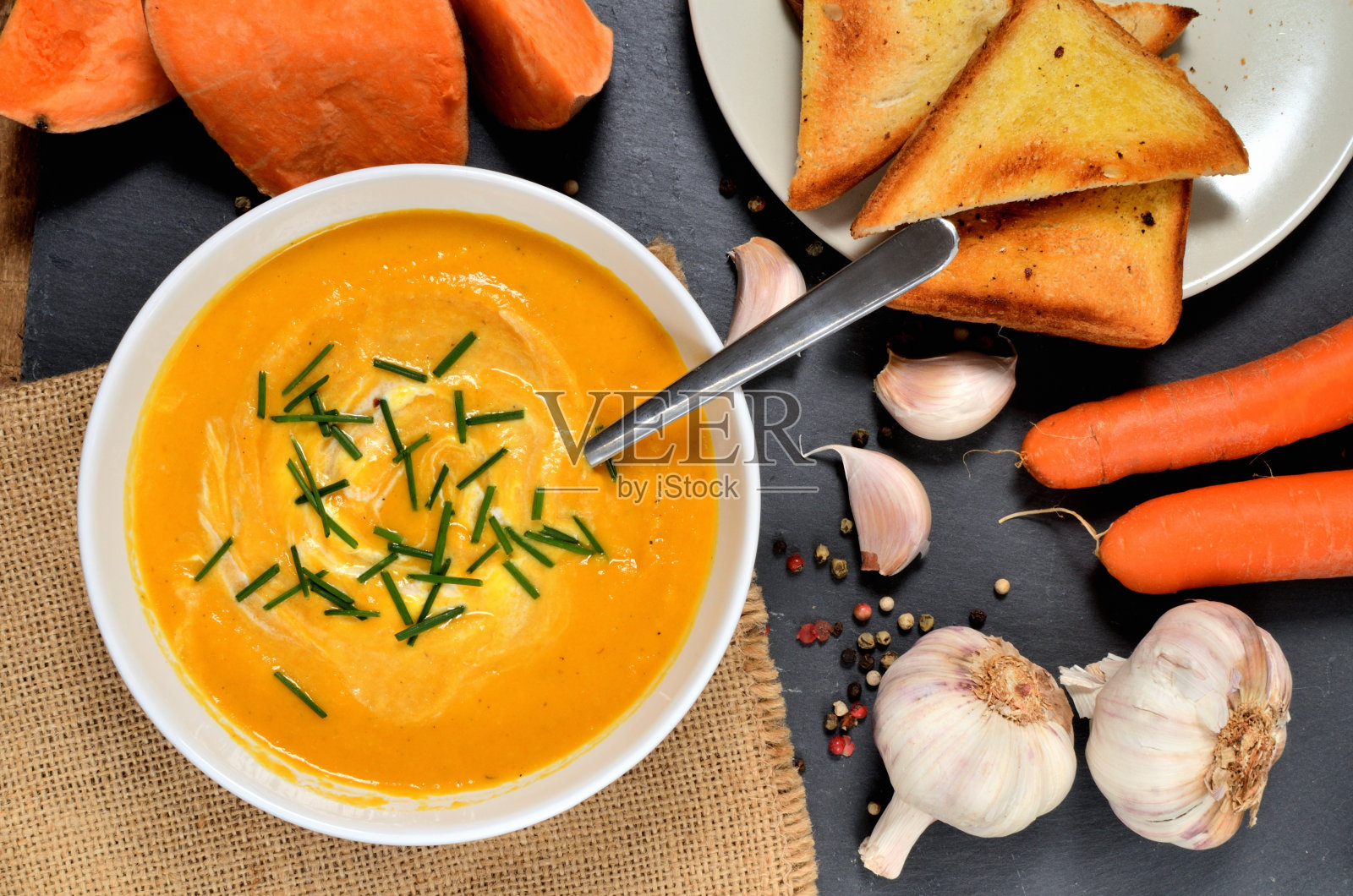 一碗甘薯奶油汤，配上鲜香葱、汤匙、胡椒、胡萝卜、大蒜和烤面包，放在石板上。前视图。照片摄影图片