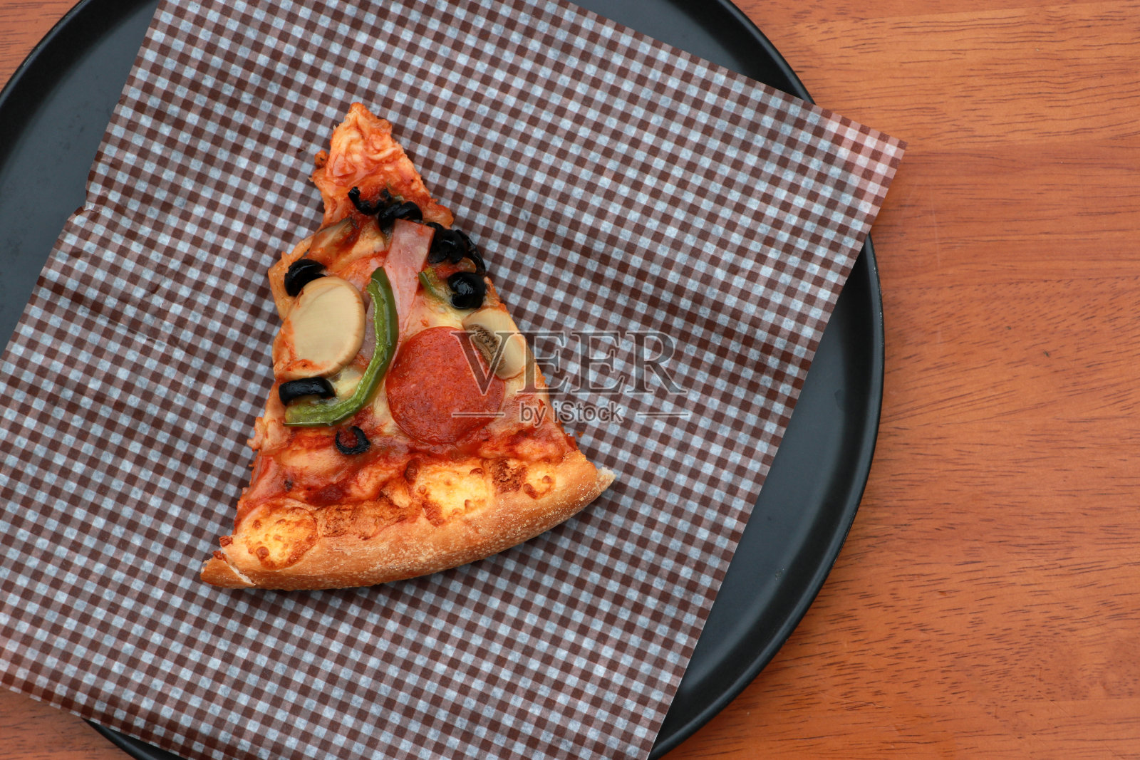 一块三角形披萨在棕色透明纸上放置在黑色圆形盘子。照片摄影图片