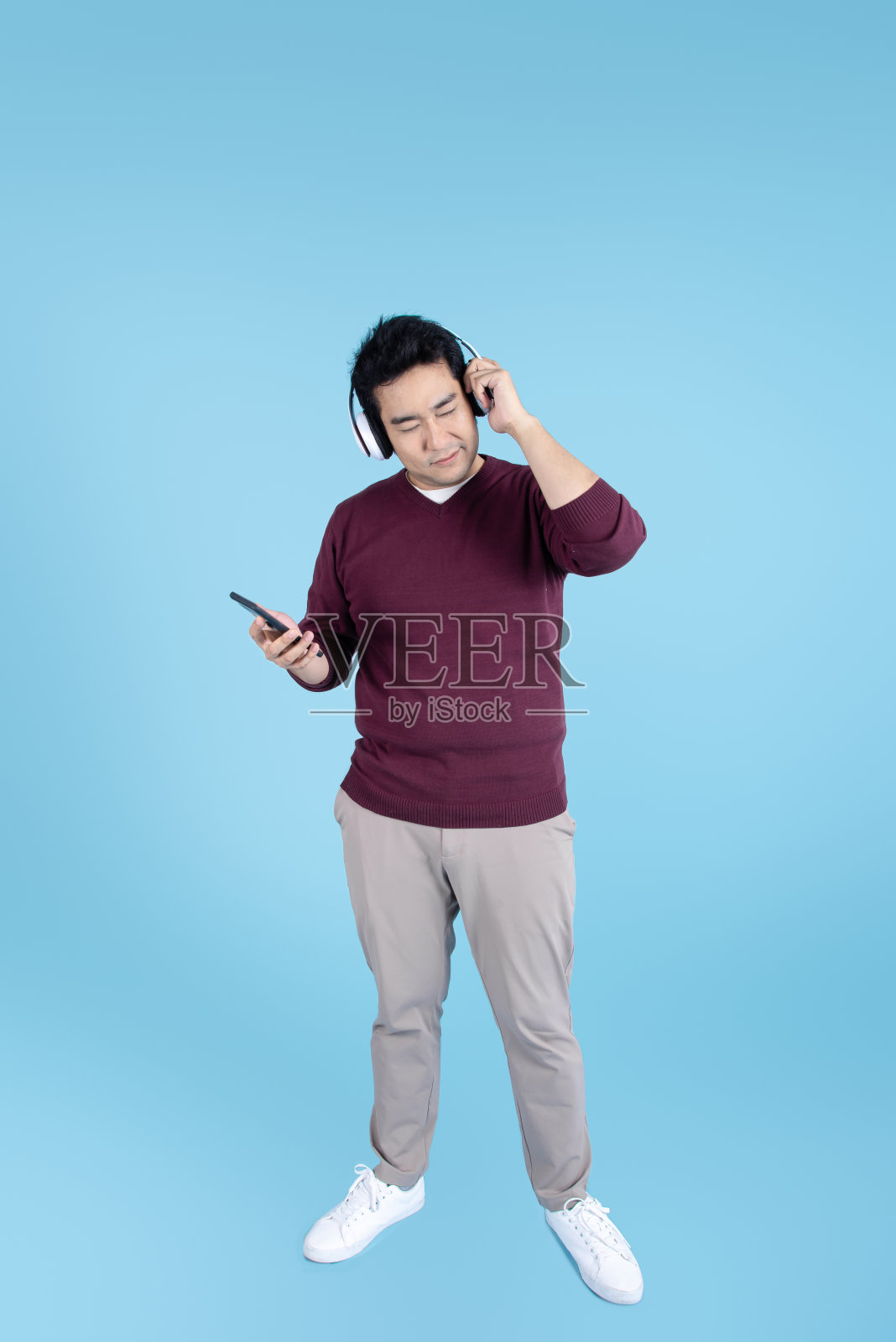 快乐的亚洲男人穿着毛衣，用耳机和智能手机在蓝色背景。照片摄影图片