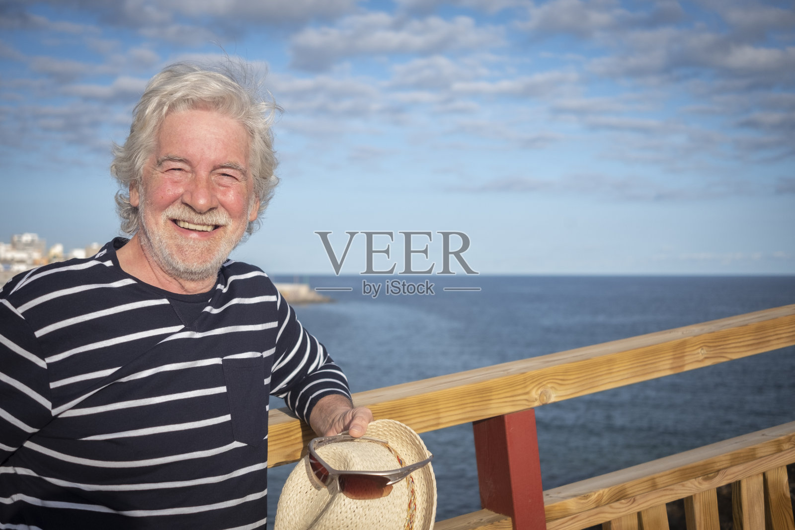 有魅力的快乐老人戴着草帽和蓝色条纹衬衫在海上远足，靠在木栈道的栏杆，蓝天和云彩的背景-积极退休老人的概念照片摄影图片