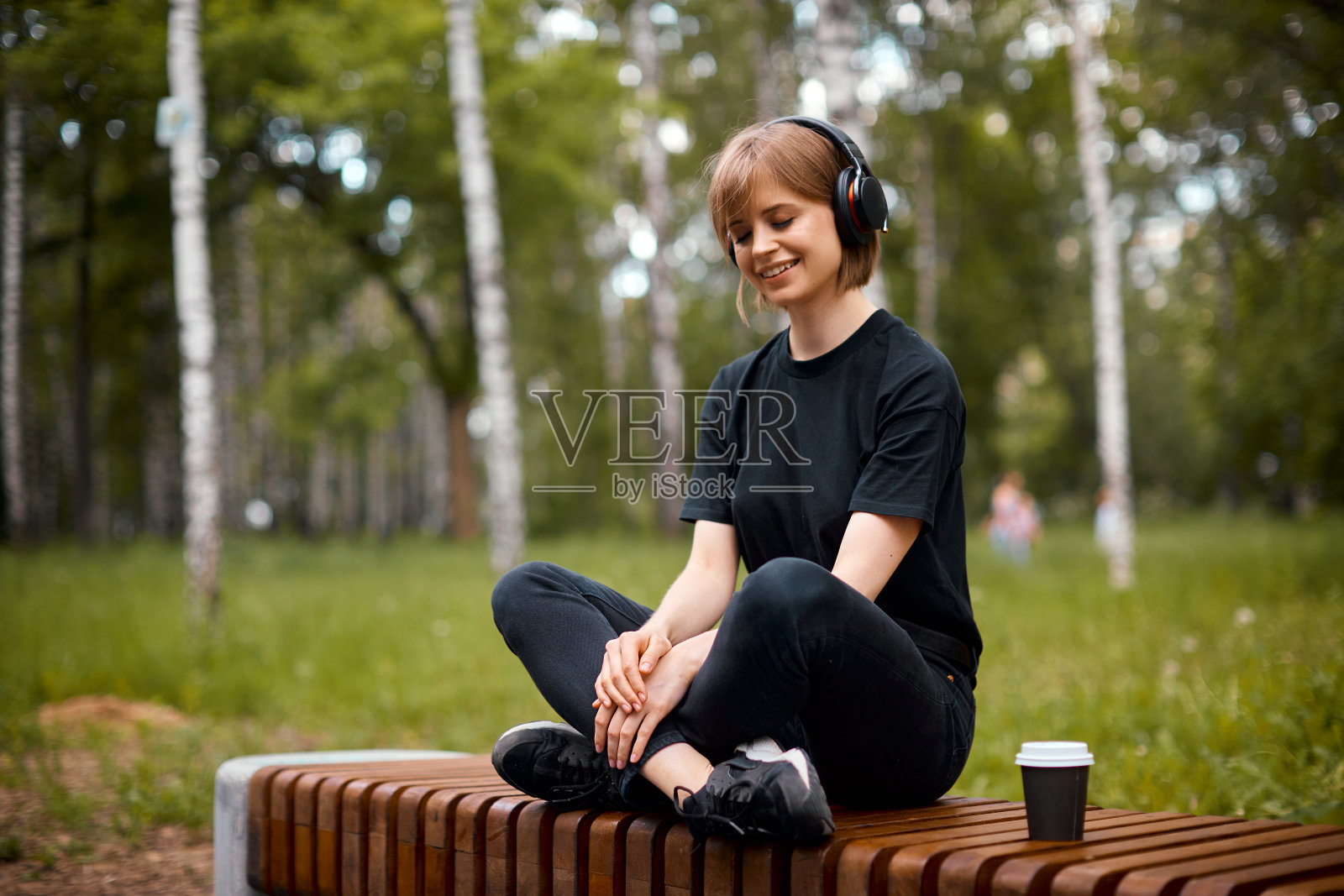 年轻女孩面带微笑，戴着无线耳机听着音乐，在公园里喝着茶，坐着莲花坐冥想照片摄影图片