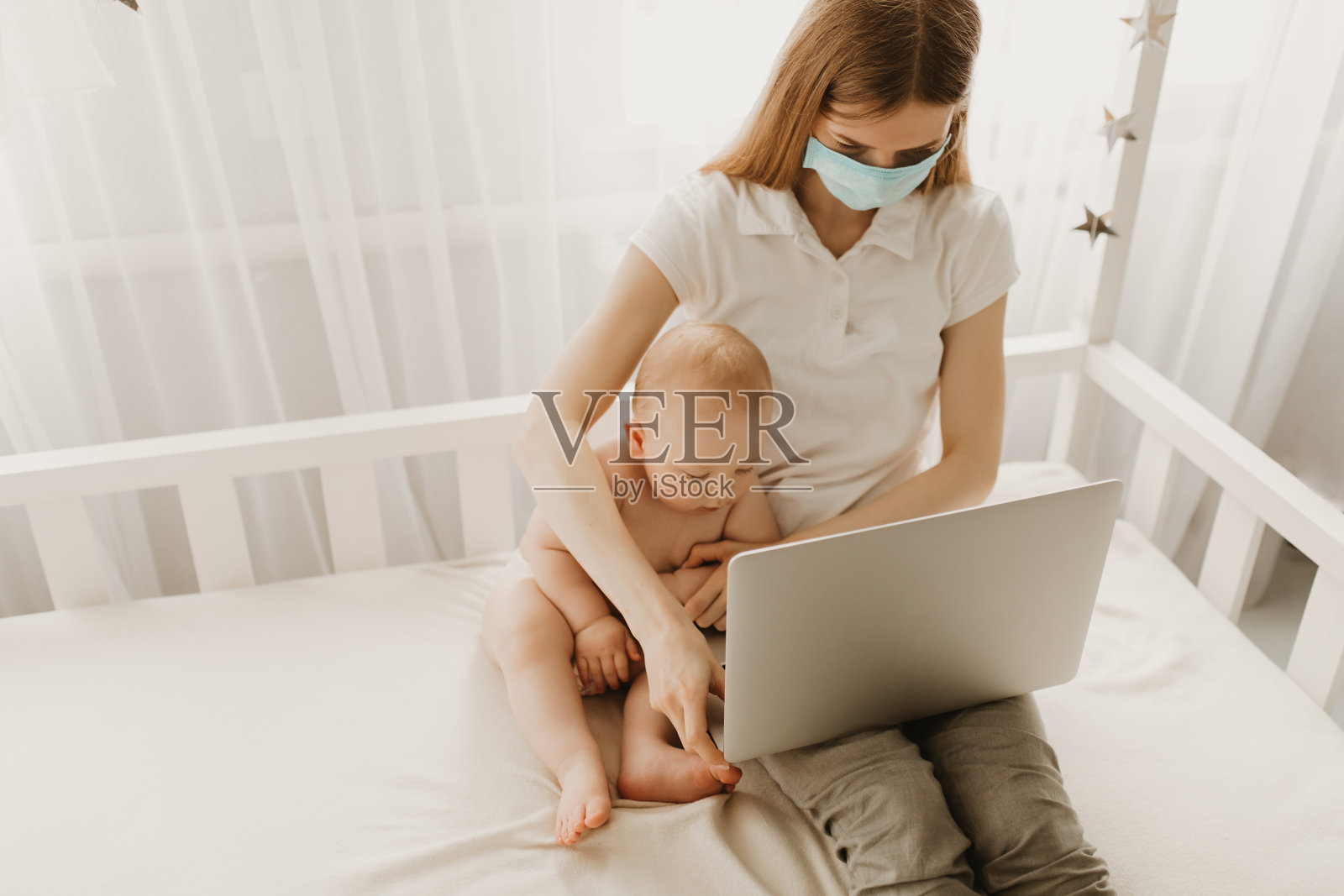 一位戴着医用防护口罩的母亲正在用笔记本电脑远程工作，一位休产假的年轻母亲试图带着孩子做自由职业。检疫、冠状病毒照片摄影图片