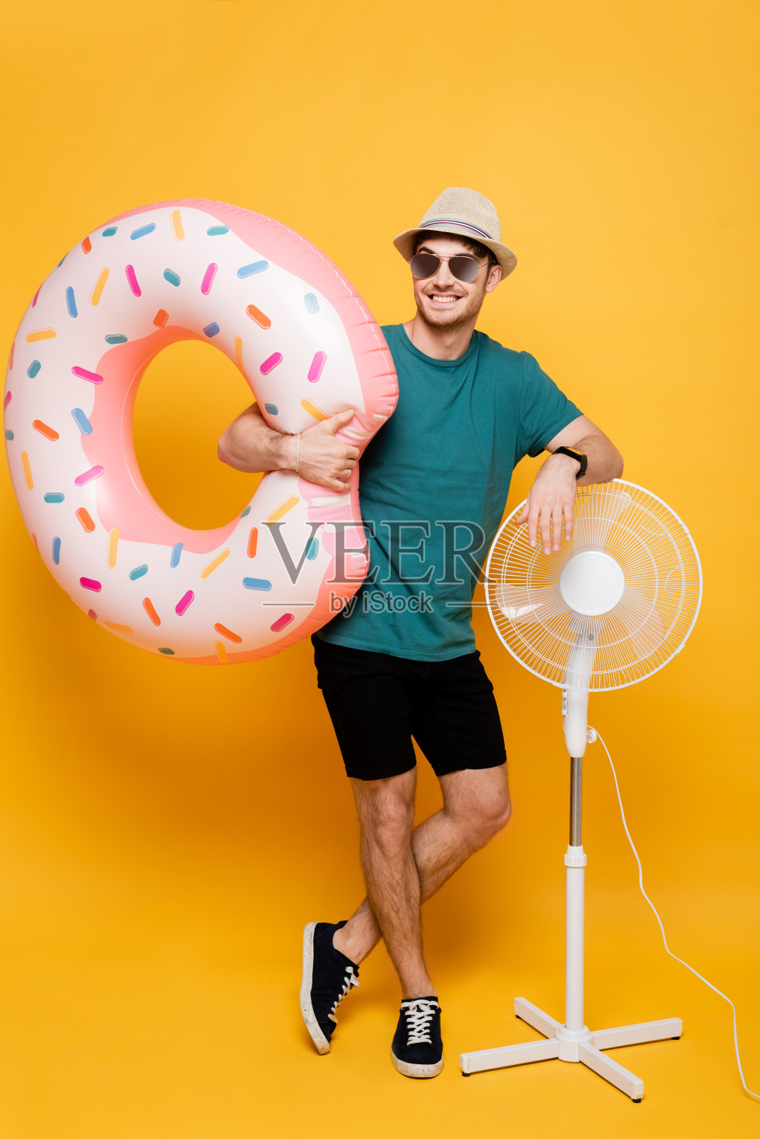 面带微笑的男人戴着墨镜和帽子，手里拿着充气甜甜圈，旁边站着黄色的电扇照片摄影图片
