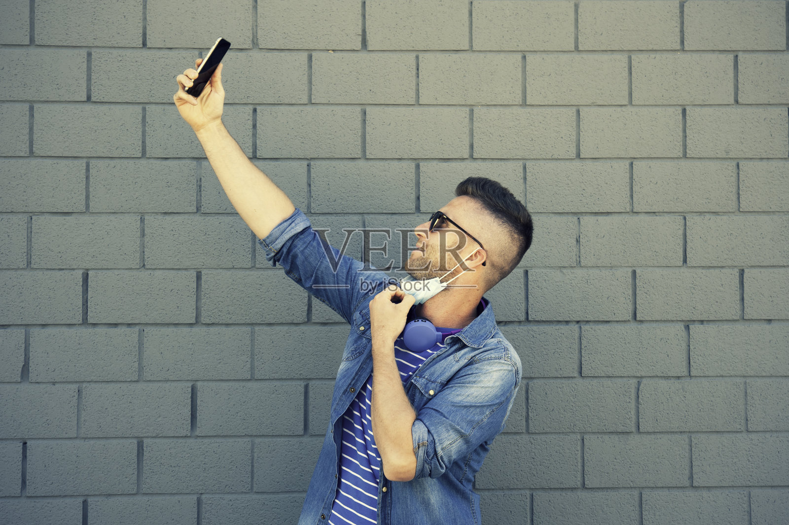 冠状病毒爆发后，年轻人戴着耳机戴着口罩——用智能手机自拍——封锁后的新常态——社交网络成瘾照片摄影图片