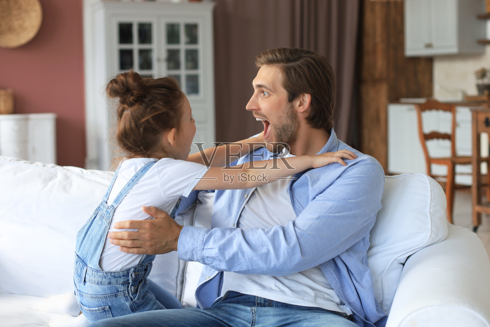 微笑的年轻父亲躺在客厅的沙发上和快乐的小女儿玩。照片摄影图片