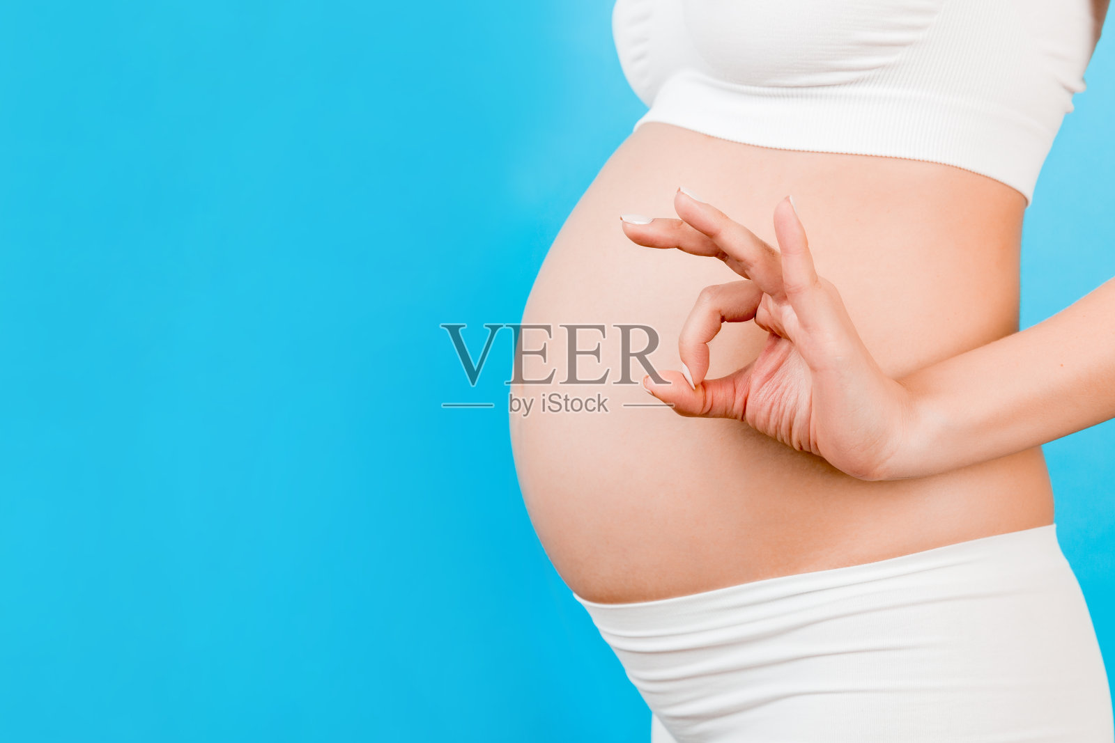 幸福的孕妇在白色内衣显示好的手势在她的腹部在蓝色背景的特写。积极的怀孕。本空间照片摄影图片