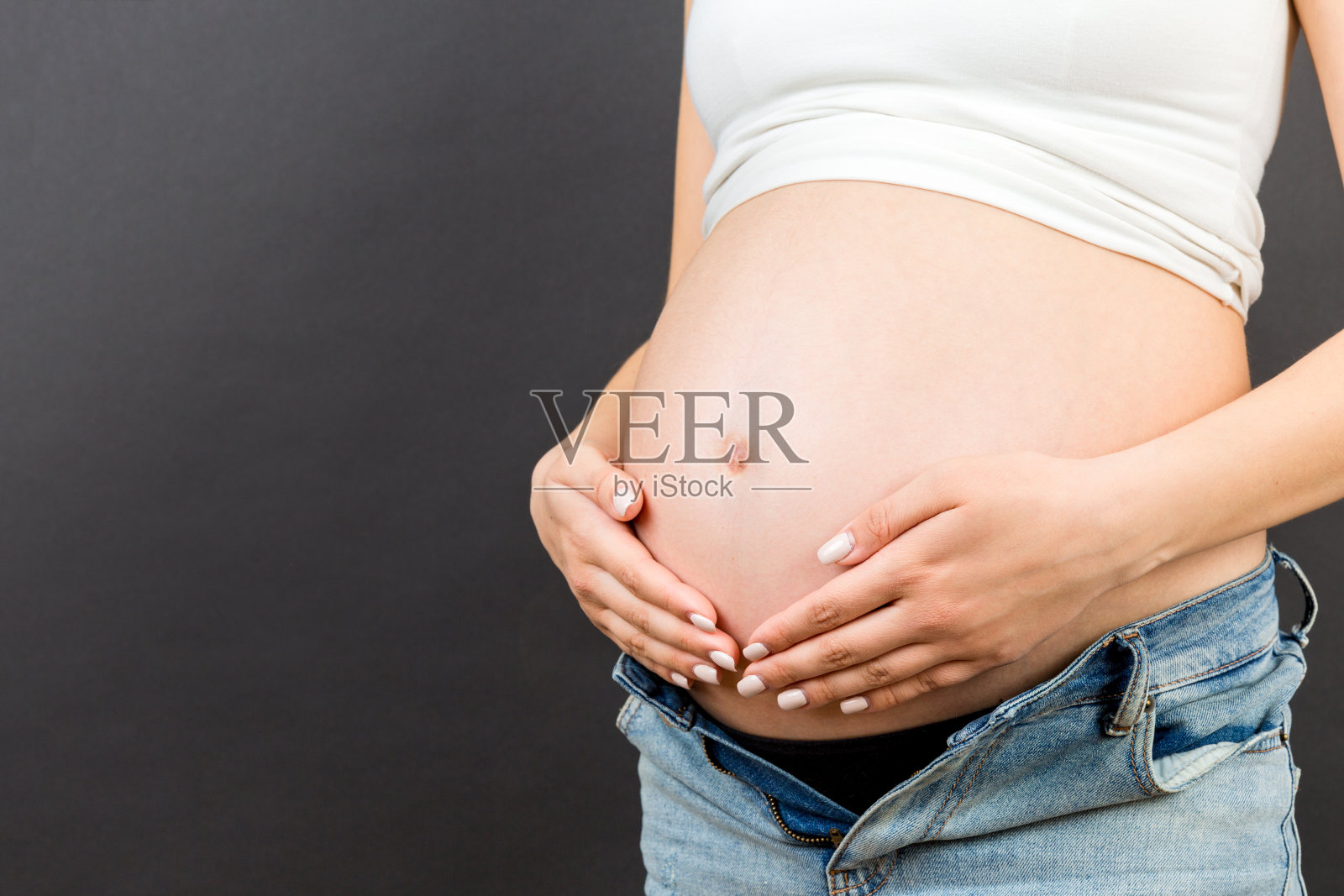 近距离的孕妇在无拉链的牛仔裤显示她裸露的腹部在彩色的背景与复制空间。宝宝期待概念照片摄影图片