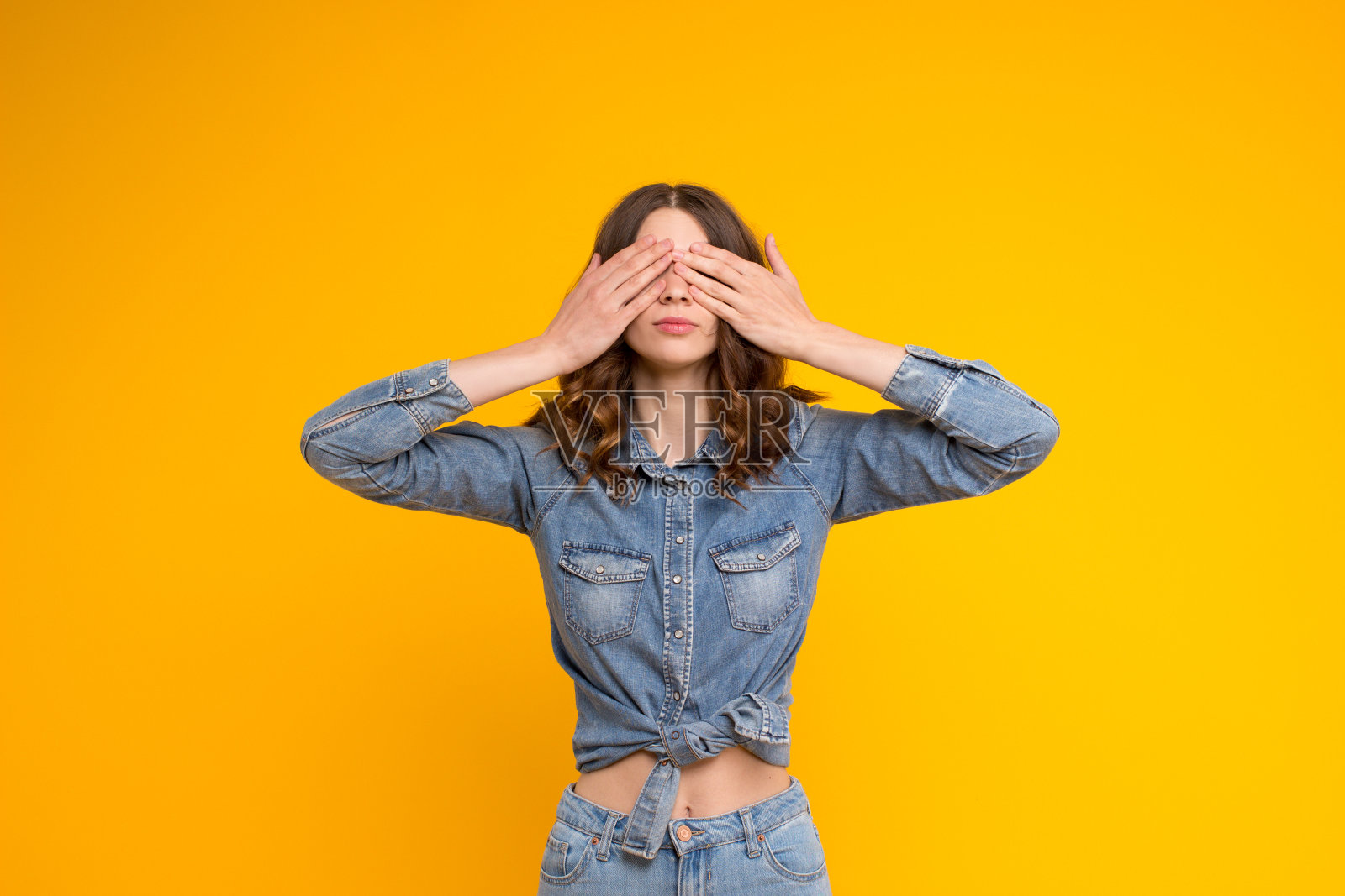 冷静美丽卷曲的黑发年轻女子在蓝色牛仔休闲衬衫和牛仔裤覆盖她的眼睛与双手，孤立地站在黄色的背景。我害怕,害羞,盲目的照片摄影图片