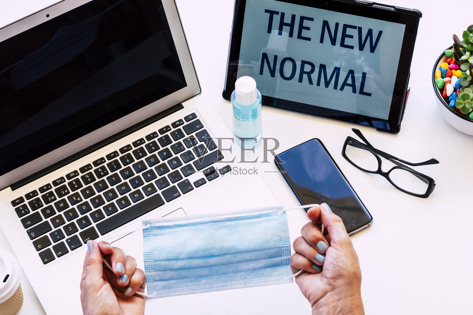 高级女性用双手捧着面膜——白色的办公桌上有笔记本电脑和带信息的平板电脑——新常态照片摄影图片