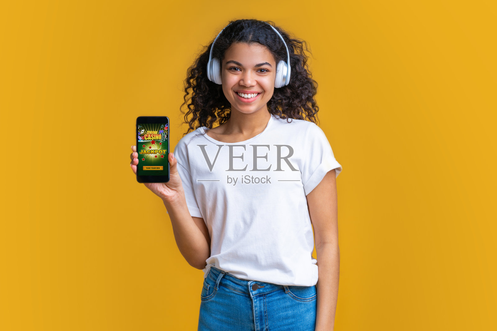 可爱的黑皮肤女孩微笑着戴着无线耳机，她的手机屏幕与博彩网站的摄像头照片摄影图片