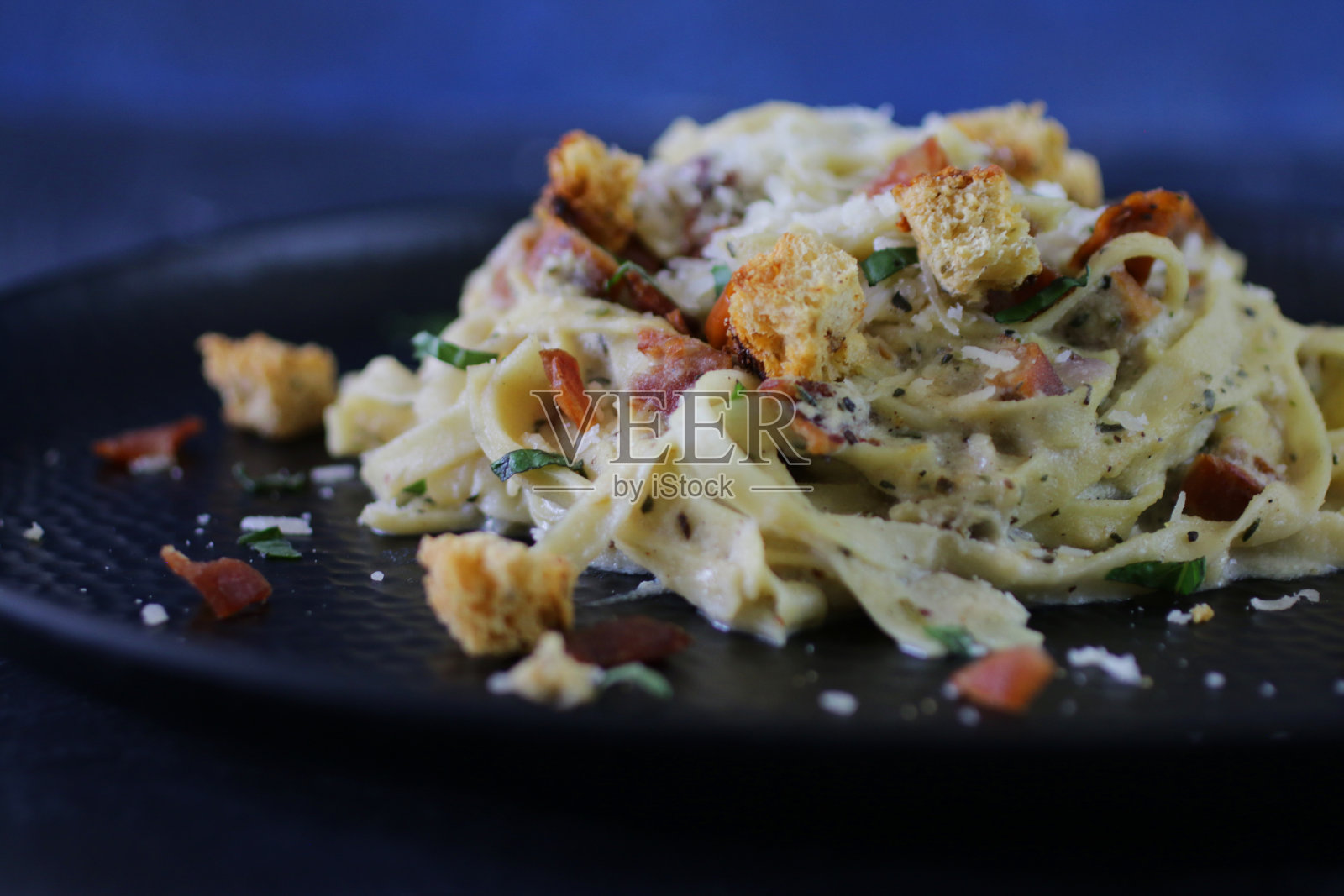这是自制意大利面加干酪沙司的特写，里面有鸡蛋、培根、意大利熏火腿和烟肉、意大利帕尔马干酪和大蒜，放在黑色的盘子里，背景是蓝色，重点放在前景照片摄影图片