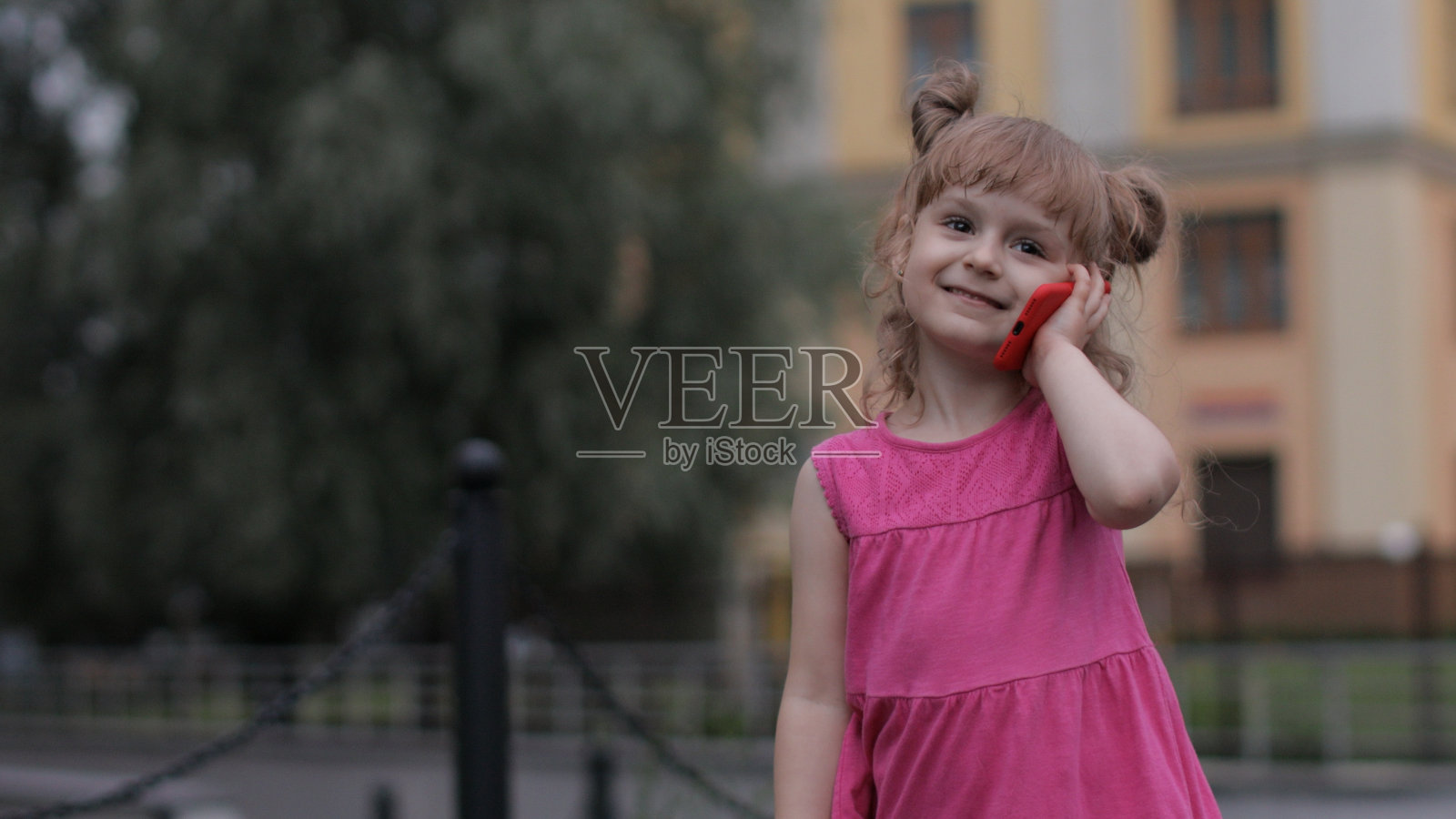 小女孩在户外用智能手机说话。穿着粉红色衣服的孩子在城市的街道上打电话照片摄影图片