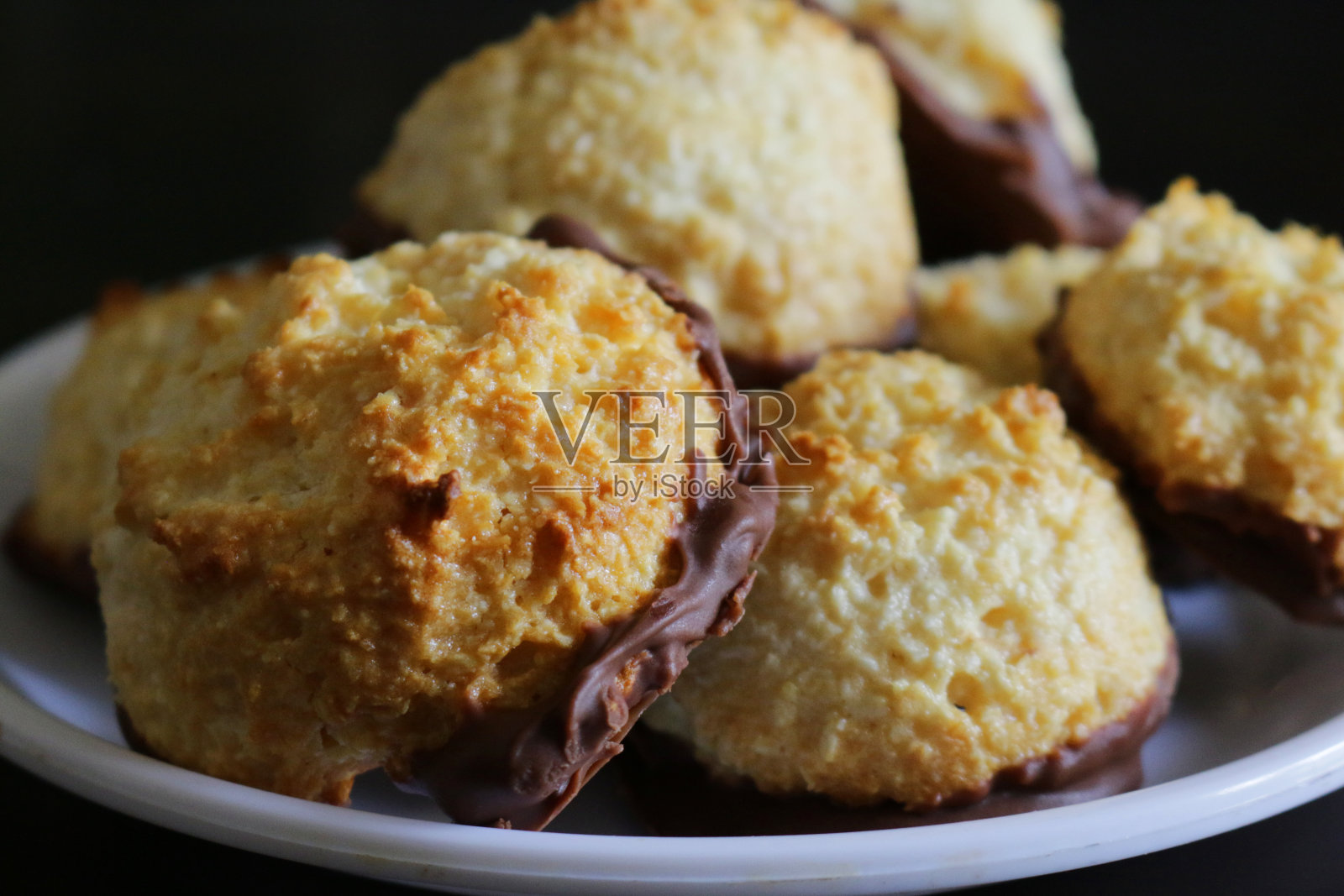 鲜烤的浸有牛奶巧克力的椰子蛋白杏仁饼堆在白色盘子上，用干椰子混合蛋白、糖、黄油和香草味制成，饼干/饼干浸在融化的巧克力中，注意前景照片摄影图片