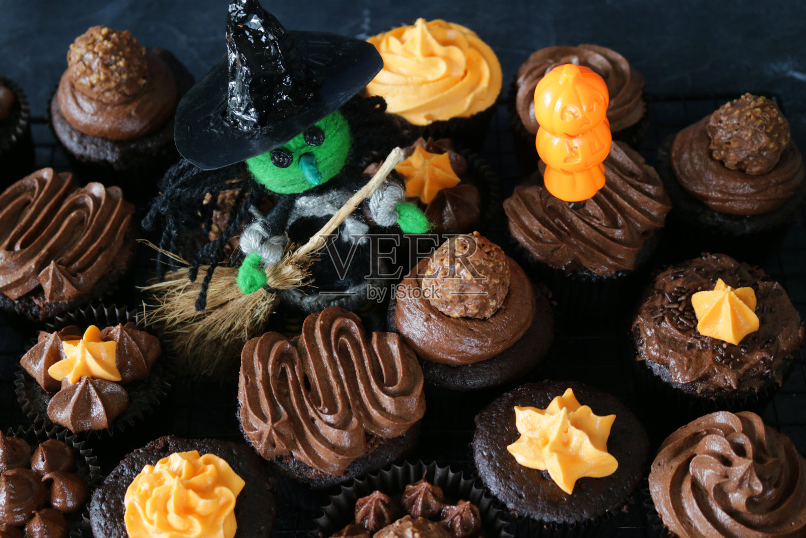 这是万圣节女巫和扫帚的特写，上面是自制的巧克力纸杯蛋糕，上面是棕色和橙色的管道糖霜，黑色的纸蛋糕盒在板石背景上，俯瞰照片摄影图片