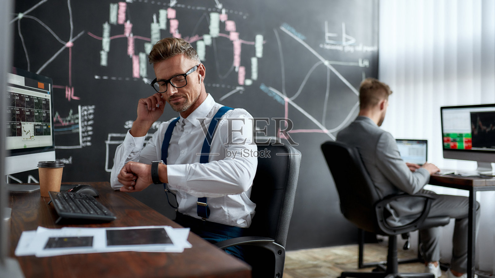 获得正确的。时尚商人，商人一边看手表一边用无线耳机工作。他的同事和黑板上满是图表和数据分析的背景。照片摄影图片