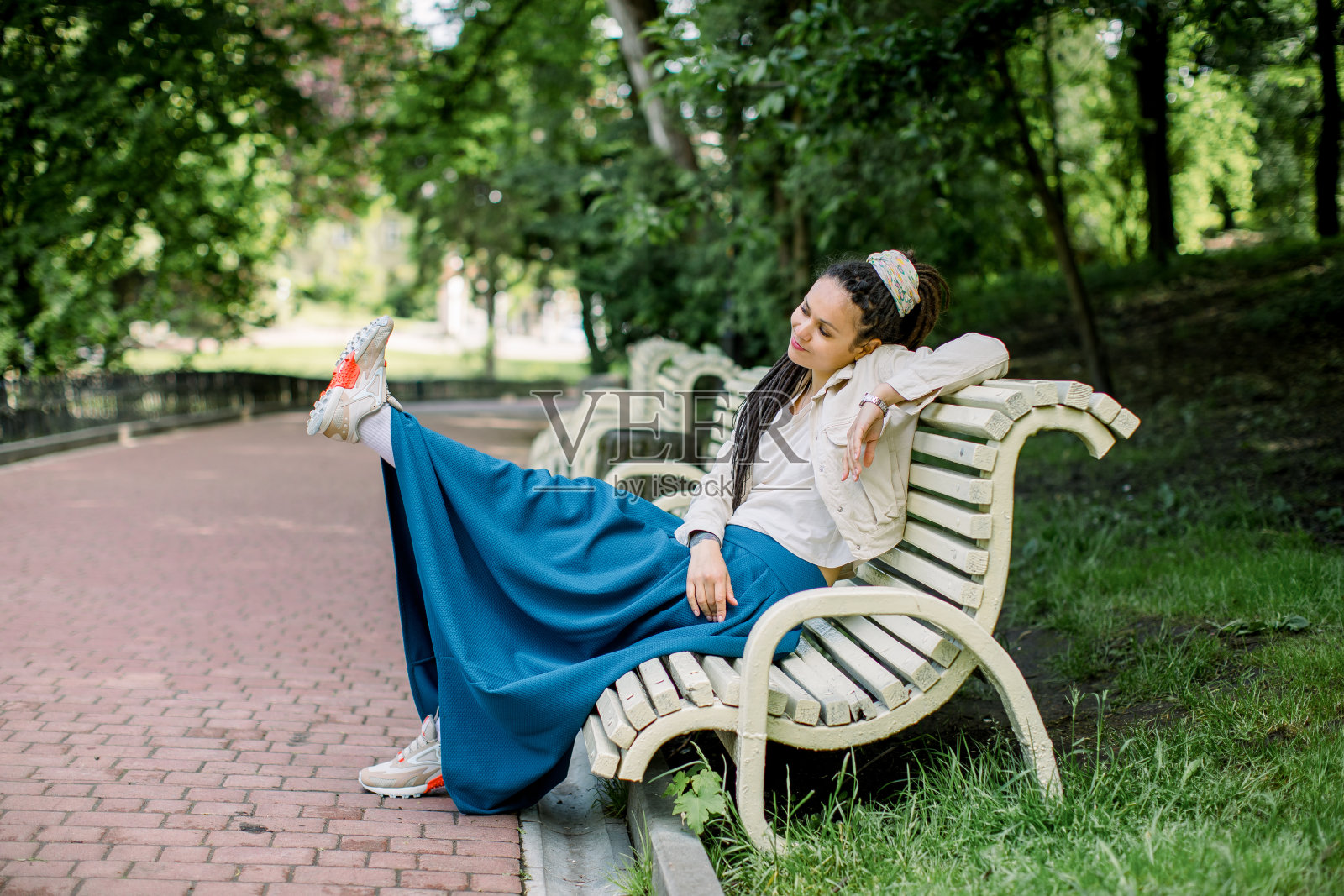 户外肖像的现代潮人女孩长发绺穿着蓝色的裙子和白色的t恤，坐在一个城市公园的长椅上，一条腿抬高和有乐趣照片摄影图片