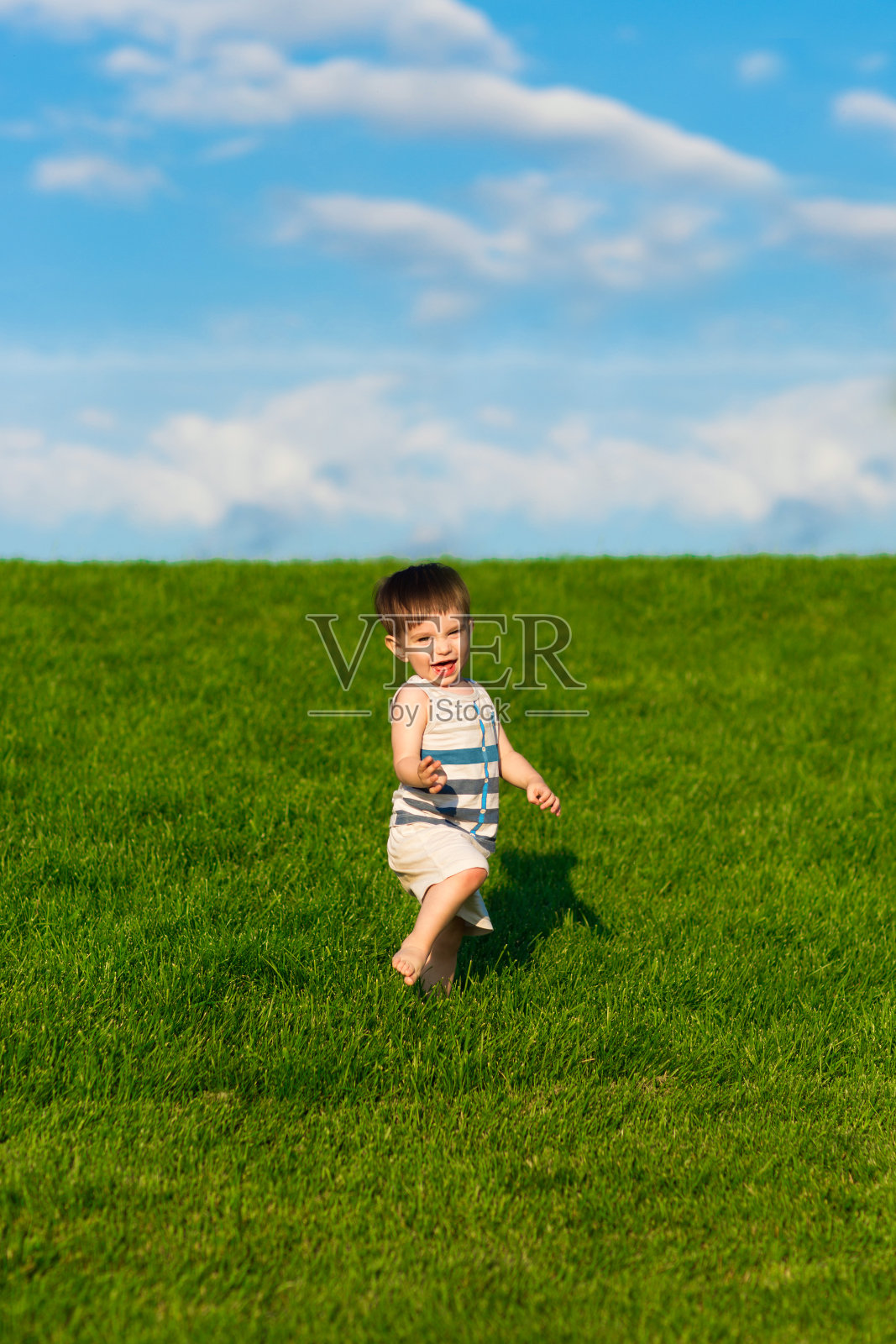 快乐的蹒跚学步的孩子在草坪上奔跑，明亮的绿色草地和蓝色的天空照片摄影图片