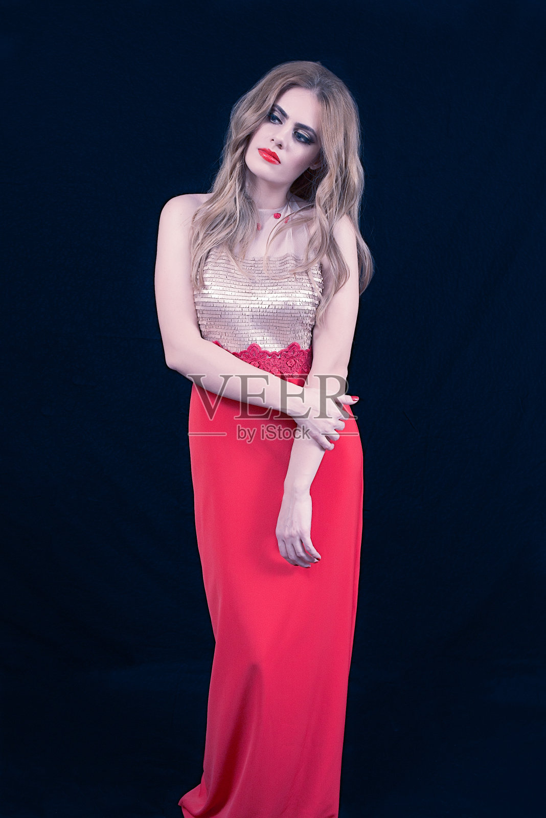 年轻的金发与金色和红色的连衣裙，保持她的手臂在一个防御的位置和看别处，在深蓝色的背景。时尚的位置照片摄影图片