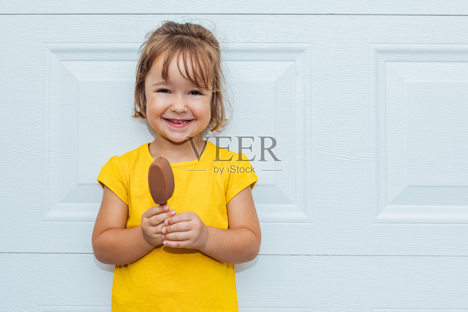 可爱的金发女孩吃着冰淇淋，穿着黄色的衬衫倚靠在白色的背景照片摄影图片