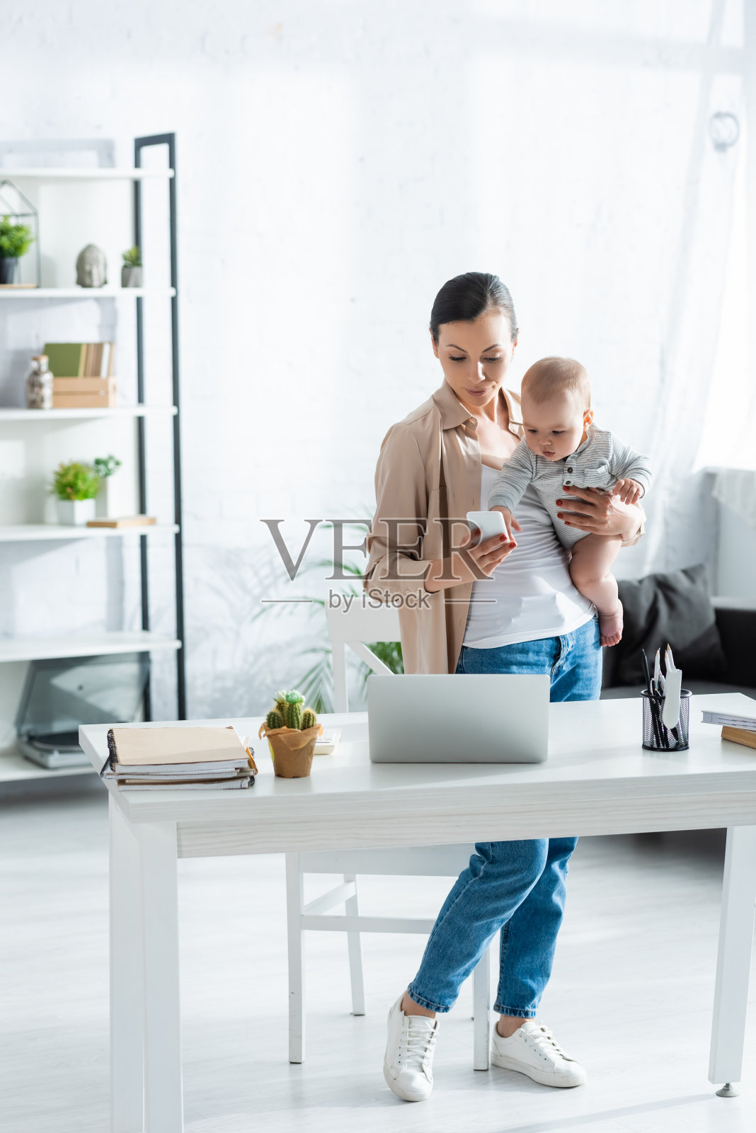 抱着婴儿的儿子，在笔记本电脑旁边使用智能手机的自由职业者照片摄影图片