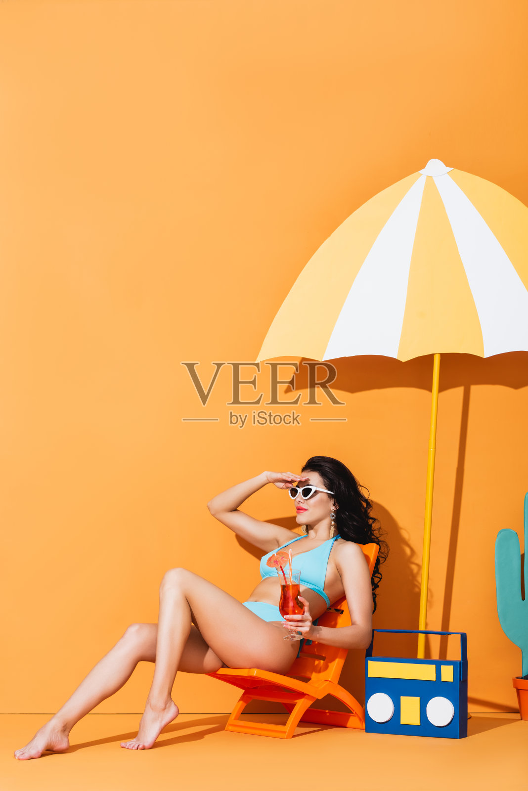 时尚的女人戴着太阳镜和泳衣坐在躺椅上，旁边是纸质音响和雨伞，手里拿着橙色的鸡尾酒照片摄影图片
