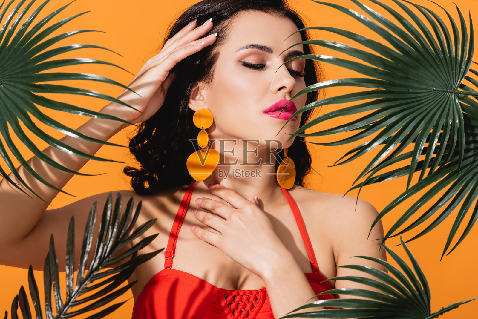 年轻女子闭着眼睛触摸脸附近的绿色棕榈叶孤立在橙色照片摄影图片