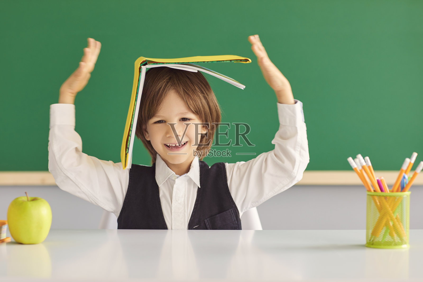 回到学校。快乐的小男孩躲在书下，在教室的课桌上玩愚蠢的游戏。照片摄影图片