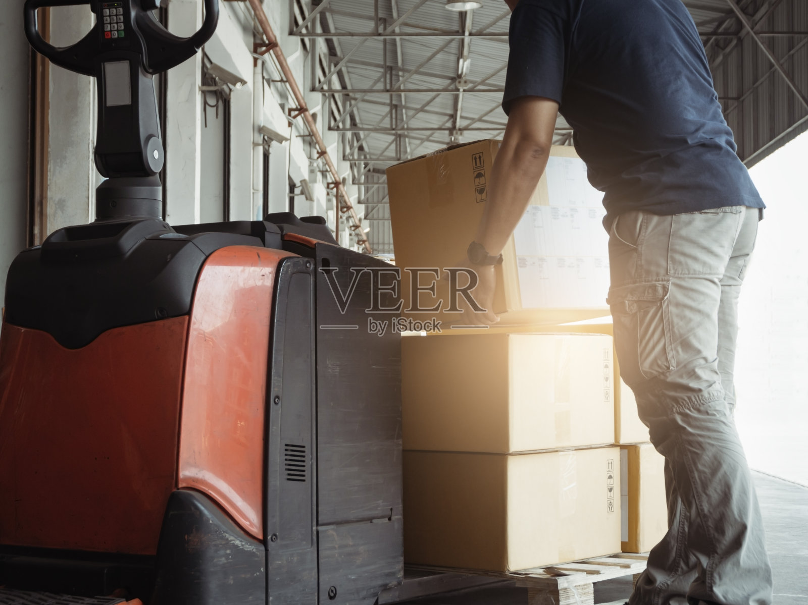 仓库工人用电动叉车千斤顶在托盘上搬运箱子。制造仓库运输包装盒。照片摄影图片