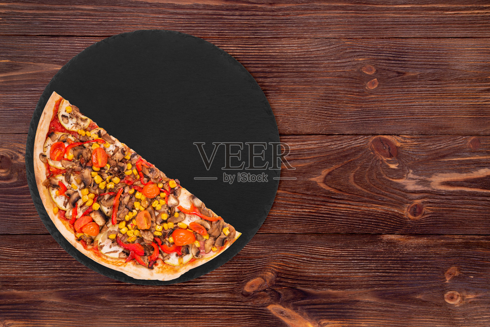 半份披萨，有蘑菇、玉米、樱桃番茄、西葫芦和甜椒，放在木桌上的石板盘子里照片摄影图片