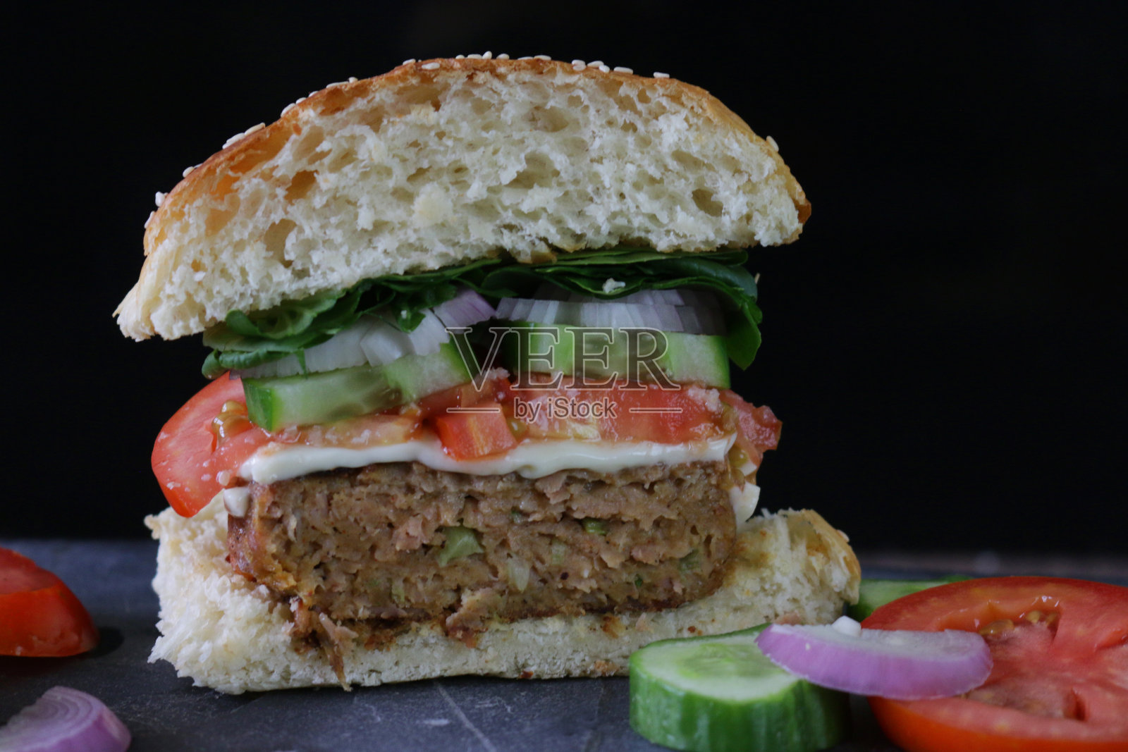 半自制的汉堡，面包包芝麻，融化的奶酪，切片的番茄，黄瓜，洋葱圈和生菜，牛肉汉堡切成两半的石板放置垫，黑色的背景照片摄影图片