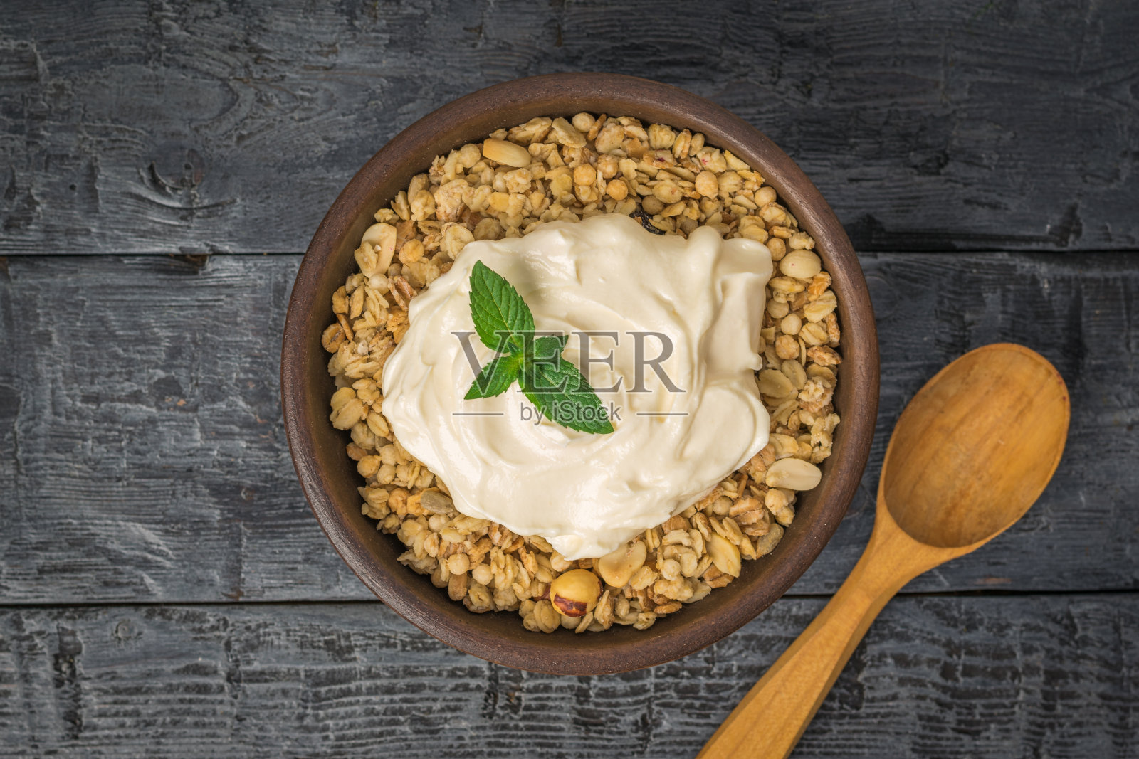 新鲜的酸奶和格兰诺拉麦片放在粘土碗里，桌子上放一个木勺。照片摄影图片