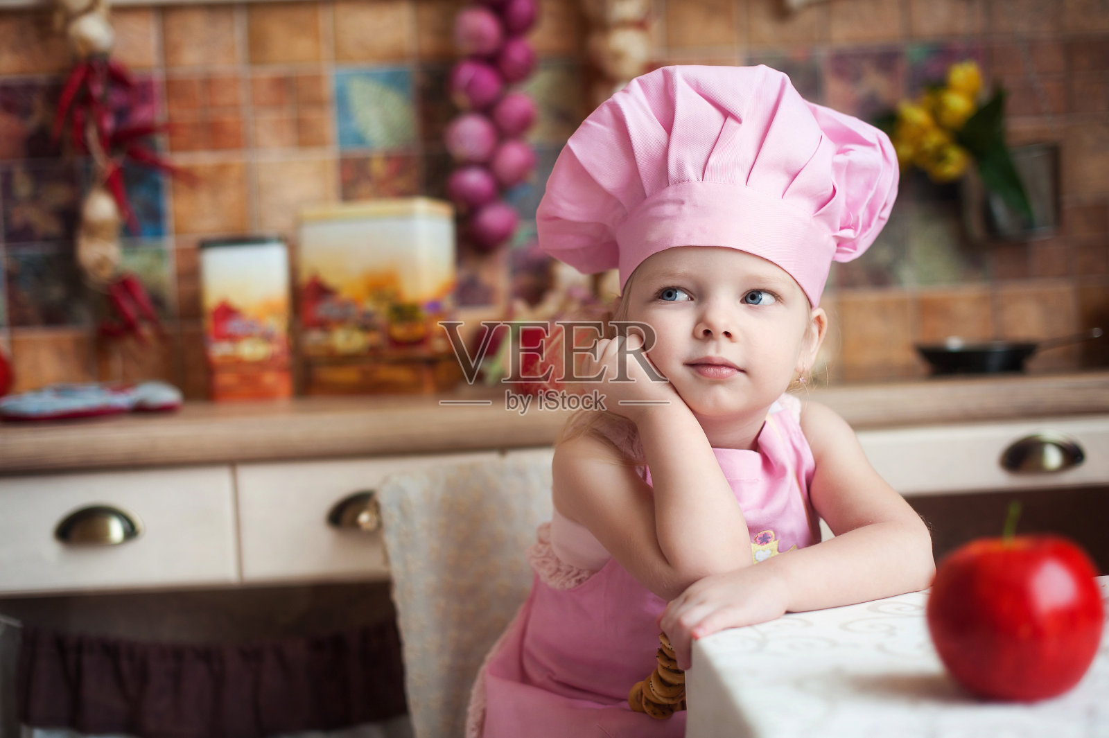厨房里，一个戴着粉色厨师帽、系着围裙的小女孩坐在桌旁，沉思着，幻想着。照片摄影图片