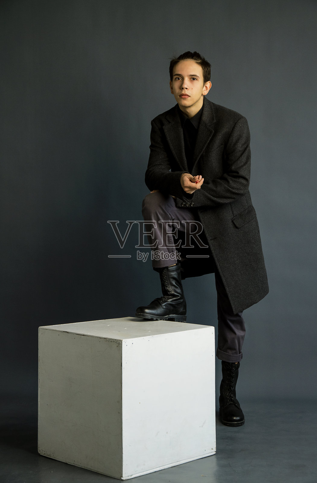 一个扎着马尾辫的年轻人，穿着经典的灰色外套和黑色的军用皮靴。在白色的立方体，灰色的背景下拍摄。照片摄影图片