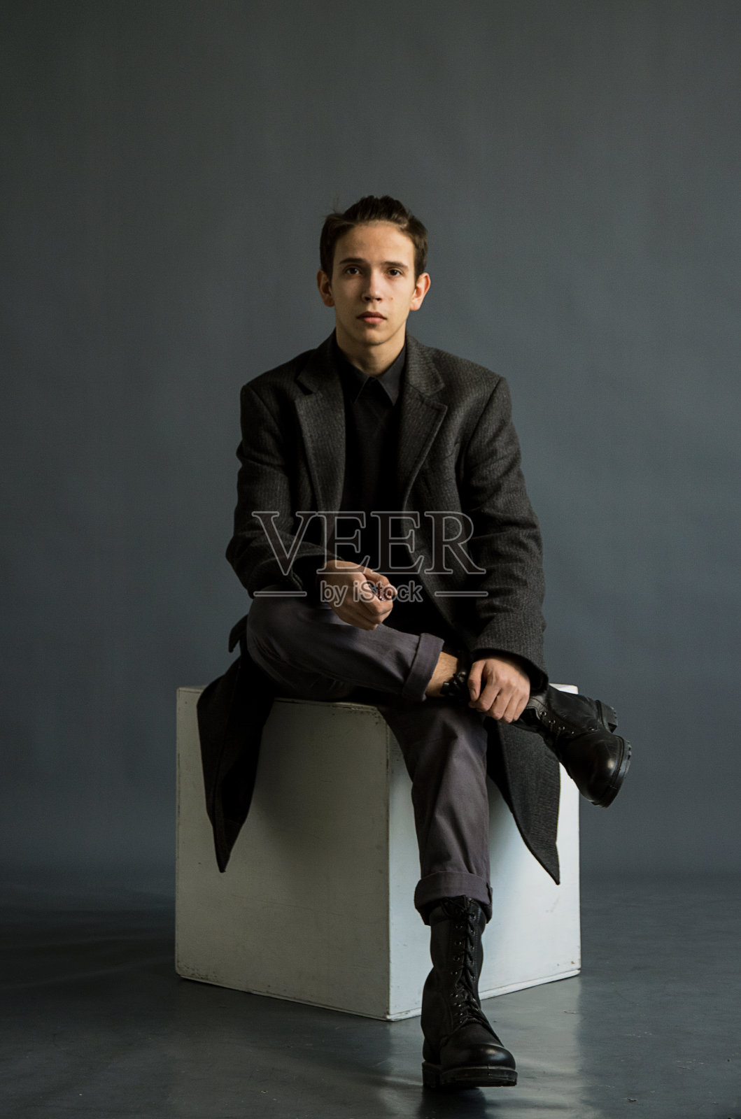 一个扎着马尾辫的年轻人，穿着经典的灰色外套和黑色的军用皮靴。在白色的立方体，灰色的背景下拍摄。照片摄影图片