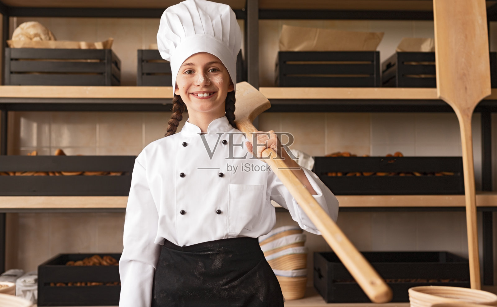 骄傲的小面包师穿着制服，拿着木器站在面包房照片摄影图片