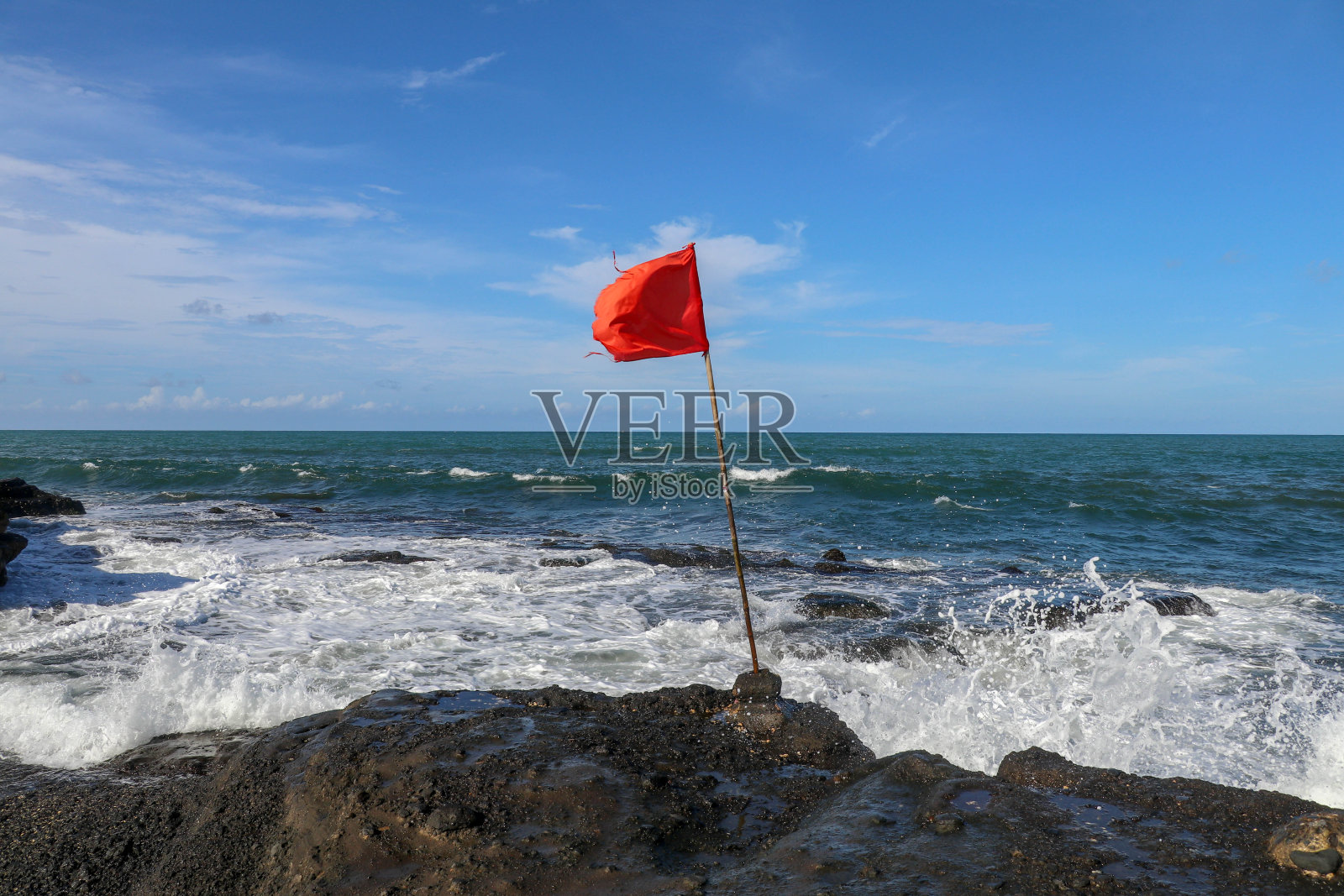 一面木制旗杆上的红旗在印度洋海岸飘扬。警告大破坏者危险的标志。在神圣的巴厘庙塔纳地段的悬崖。海浪拍打着黑色熔岩的峭壁。照片摄影图片