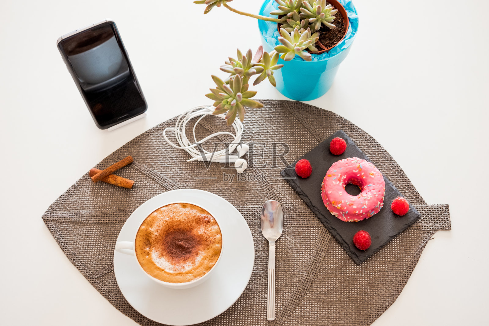 一份甜蜜的早餐，一杯热腾腾的自制卡布奇诺和一个美味的粉色甜甜圈。白色的桌上放着植物和手机照片摄影图片