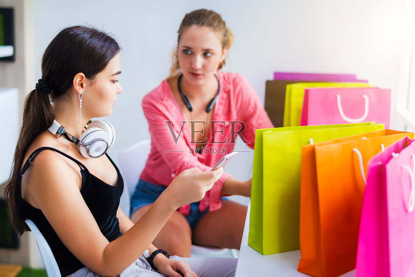 两个快乐的年轻漂亮的白人女性听音乐通过耳机连接到智能手机或手机。购物后在咖啡店或咖啡店照片摄影图片