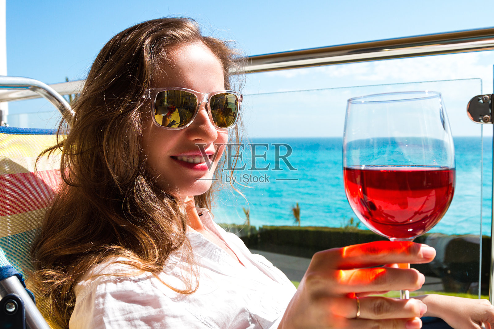 这个漂亮的长发女人戴着太阳镜，戴着玫瑰酒杯，坐在公寓的阳台上看着照相机，背景是海边的景色。照片摄影图片