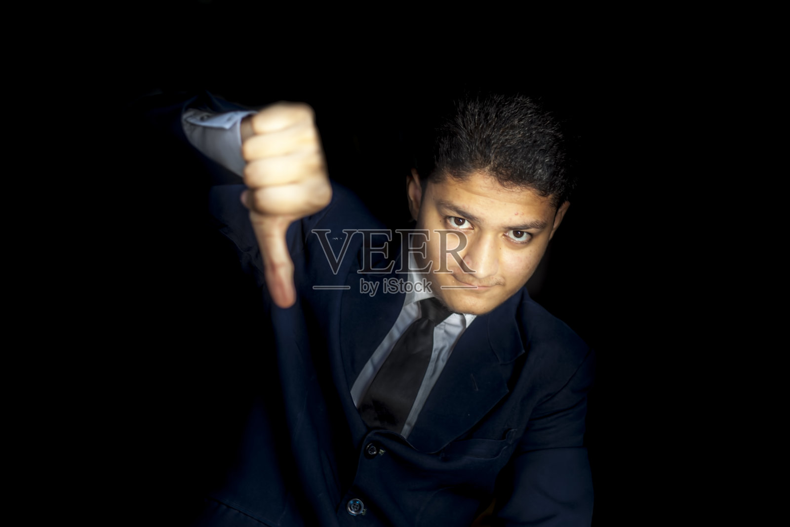 自信的商人在黑色的背景下摆出秘密姿势，深蓝色的西装上有反向的拇指符号。带着厌恶手势的男人。照片摄影图片