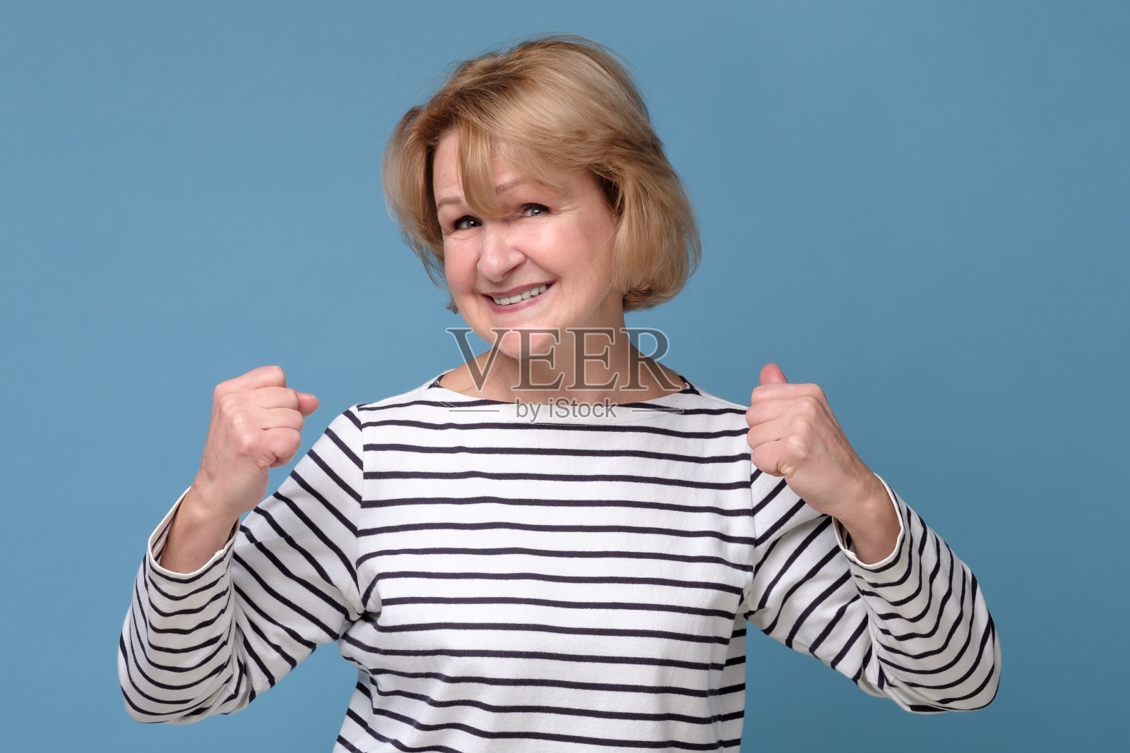 快乐的老女人举起她的拳头，孤立在蓝色的墙上。照片摄影图片