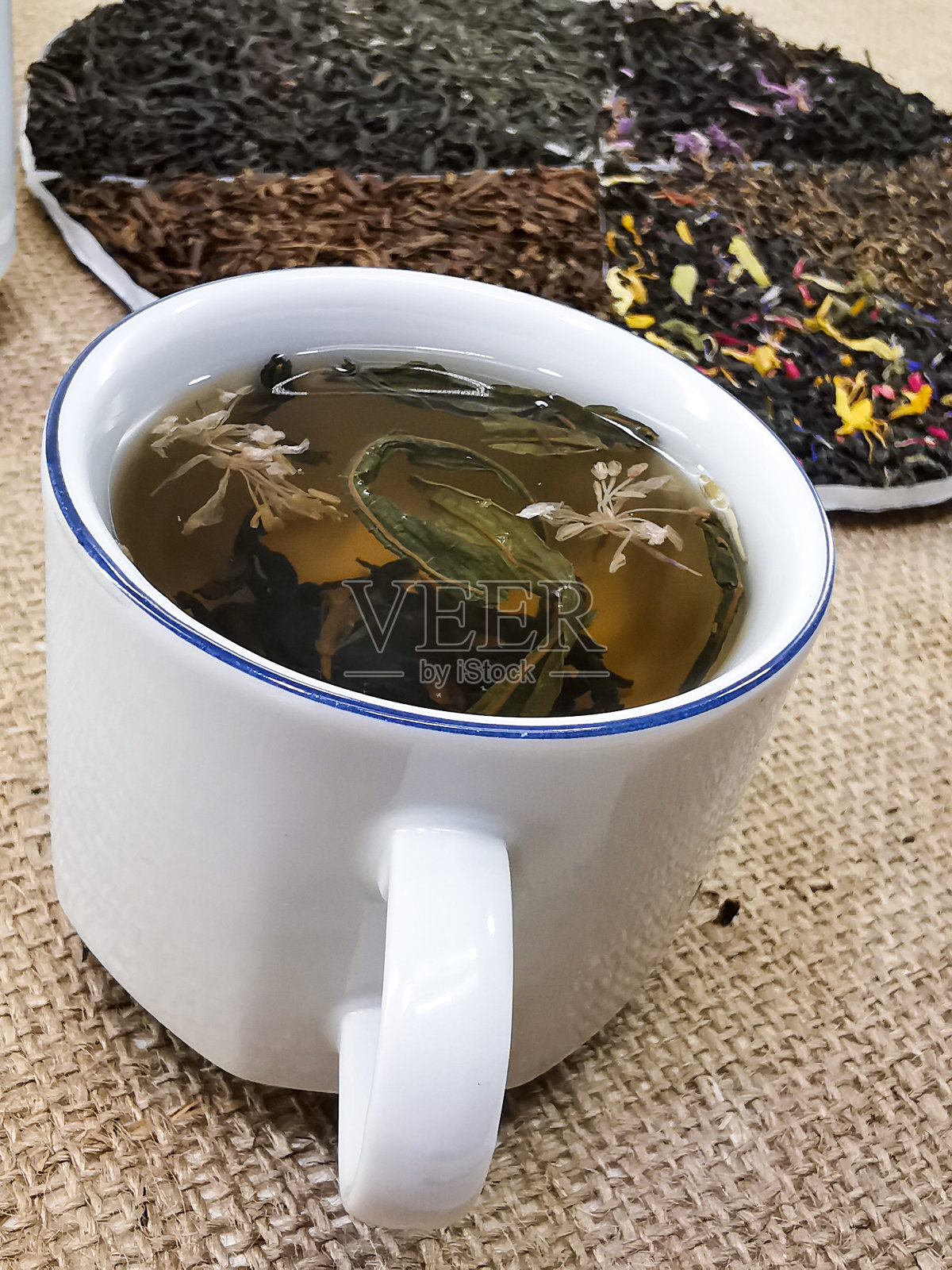 健康的生活方式的概念。将绿茶和红茶混合加入花的混合物。茶是在杯子里泡的。照片背景是粗麻布。照片摄影图片