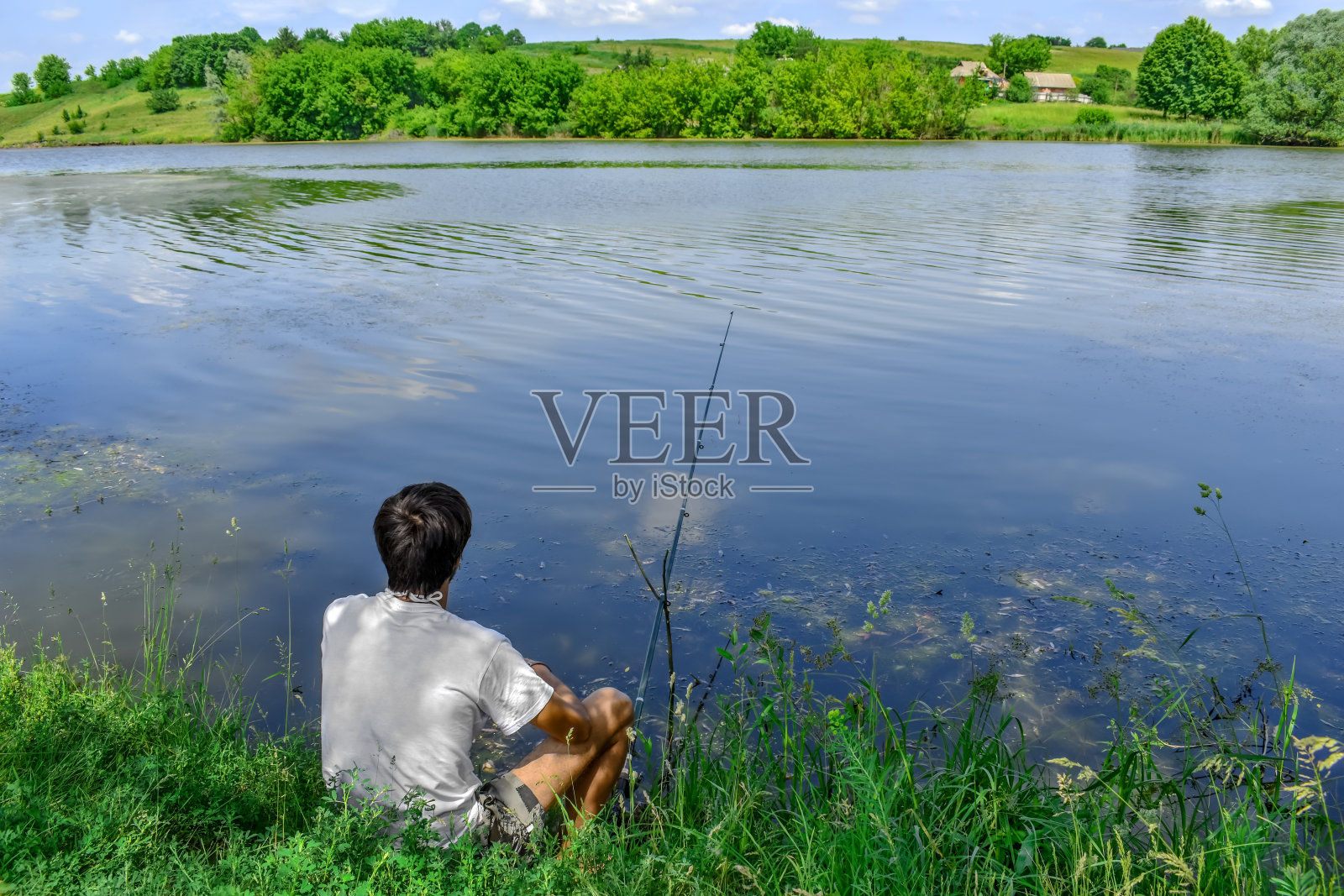 一个男人坐在池塘岸边的草地上钓鱼，背景是湖对面的乡村房屋照片摄影图片