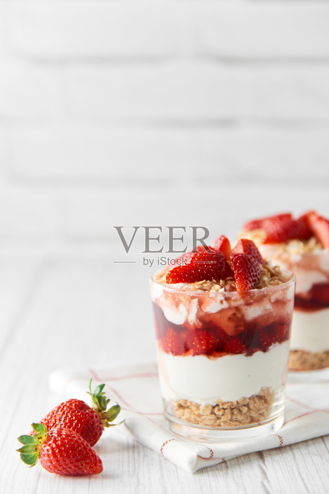 自制的分层甜点与新鲜的草莓，奶油奶酪或酸奶，格兰诺拉麦片和草莓果酱的眼镜在白木背景。健康有机早餐或零食的概念。有选择性的重点。照片摄影图片