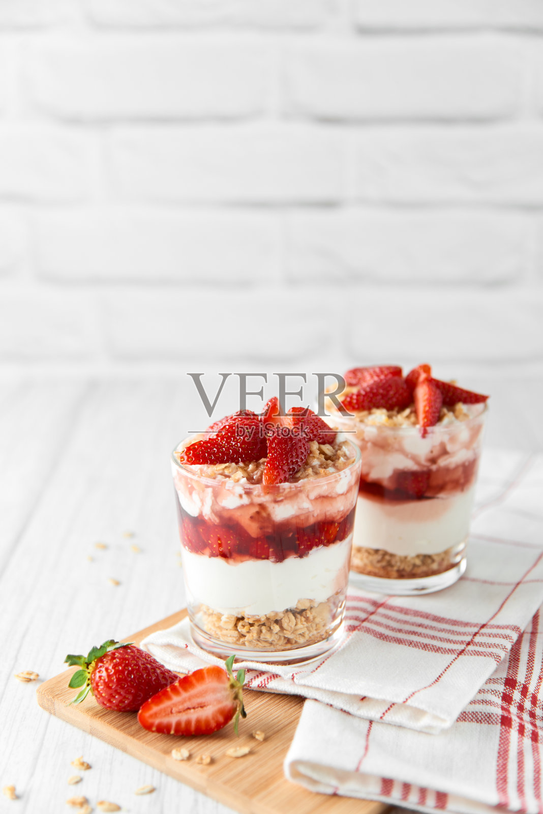 自制的分层甜点与新鲜的草莓，奶油奶酪或酸奶，格兰诺拉麦片和草莓果酱的眼镜在白木背景。健康有机早餐或零食的概念。有选择性的重点。照片摄影图片