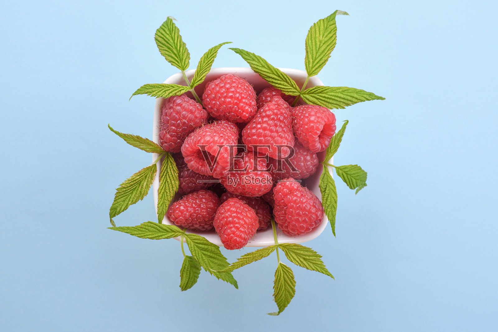 一碗新鲜的树莓，绿色的叶子，蓝色的背景。俯视图照片摄影图片