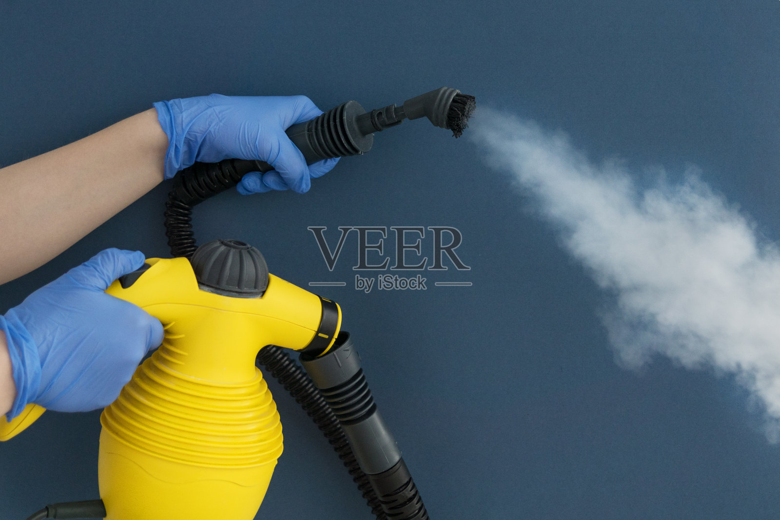 女手在蓝色手套握软管刷和家庭蒸汽发电机在蓝色背景。照片摄影图片