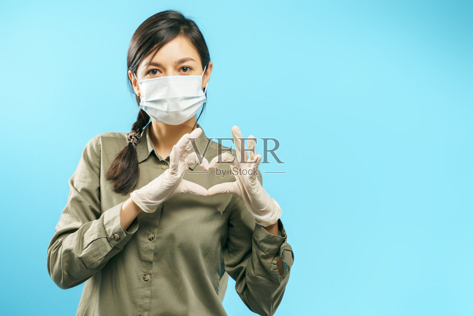 一名年轻女子的肖像与防护医疗面罩和手套显示心形手势在一个蓝色的背景。照片摄影图片